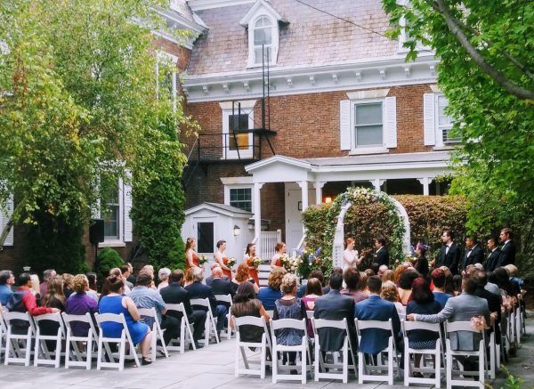 A wedding takes place in out courtyard. The bride and groom stand under a alter and rows of people water